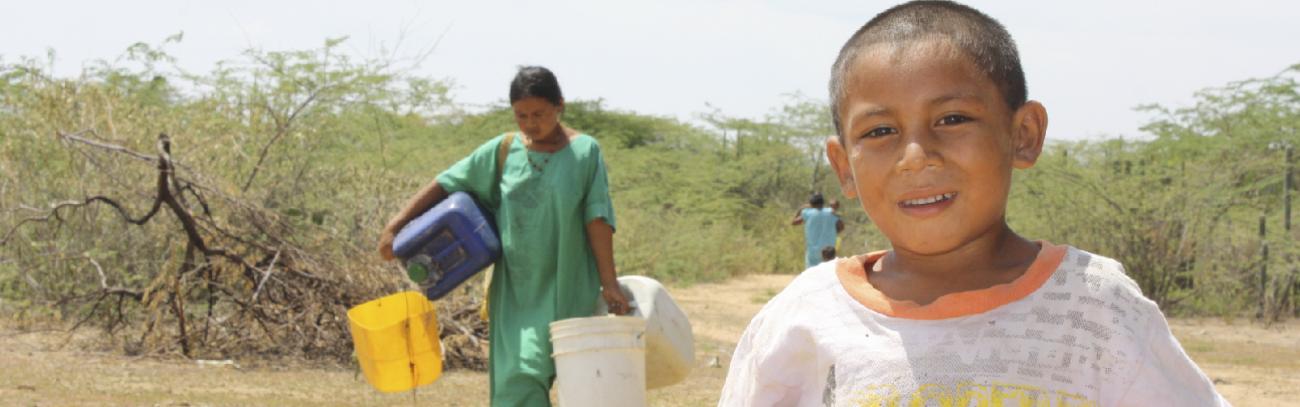 Niño en la Guajira mirando la cámara y detrás de él viene una mujer cargado 4 baldes de agua 